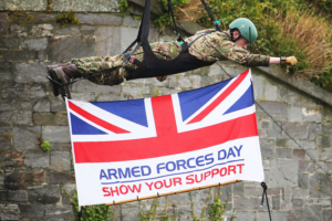 A Royal Marine zips down a zip wire with the Armed Forces flag underneath