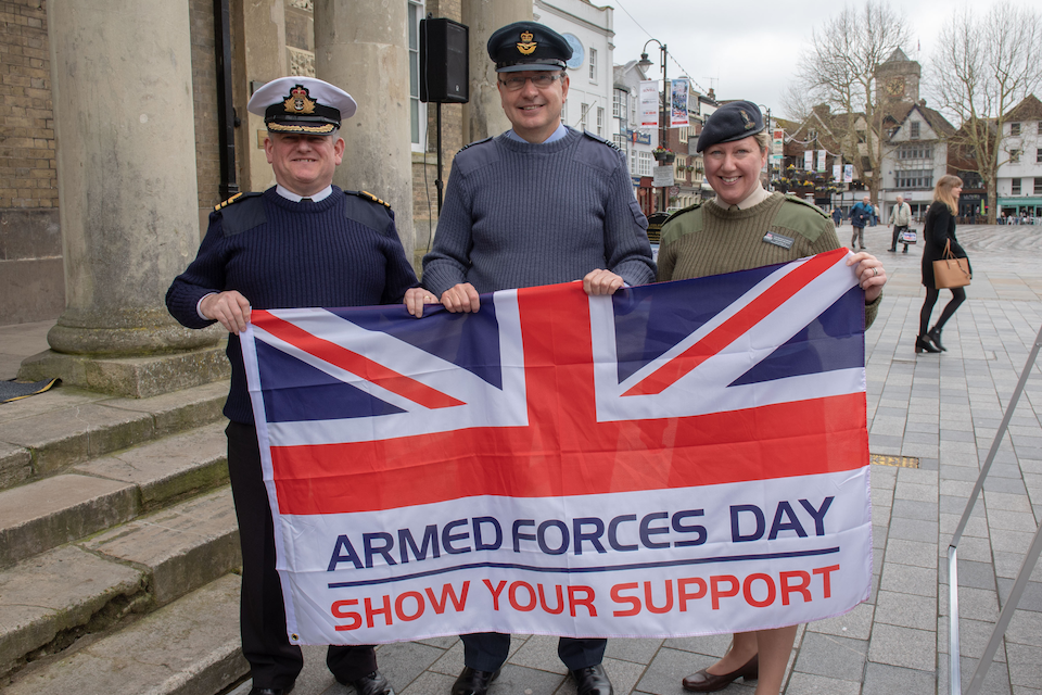 Tri-Service personnel pictured with the Armed Forces Day flag smiling. 