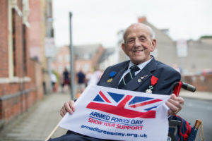 Elderly veteran wearing medals smiles at the camera holding an Armed Forces Day flag