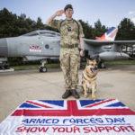 Royal Air Force Corporal stands in front of a fast jet saluting with his Service dog sitting beside him with an Armed Forces Day flag in the foreground.