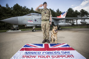 Royal Air Force Corporal stands in front of a fast jet saluting with his Service dog sitting beside him with an Armed Forces Day flag in the foreground.
